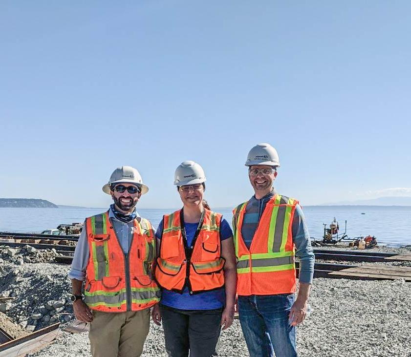 Three people in hard hats, 安全背心和安全眼镜矗立在一座正在建设的铁路桥前，背景是普吉特海湾
